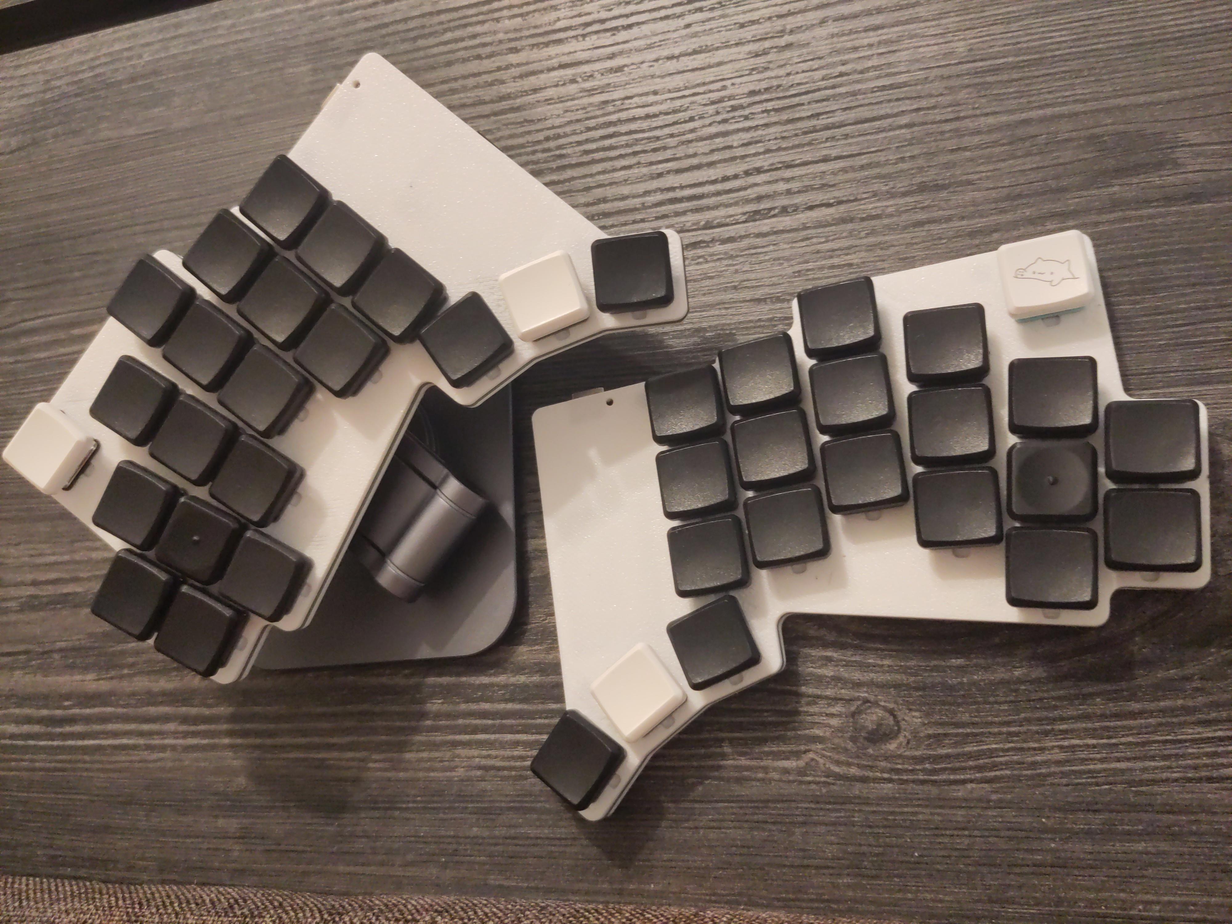 Low-profile keyboard with white casing and mostly black keycaps. The top outer corners have keys that sit a bit apart from the rest of the keys. They are white, and the one on the right half has Bongo Cat printed on it. The home keys of the pinkies have a nub on them. The home keys of the thumbs are white. To better display it, the left half is slightly raised using a magnetic phone holder.